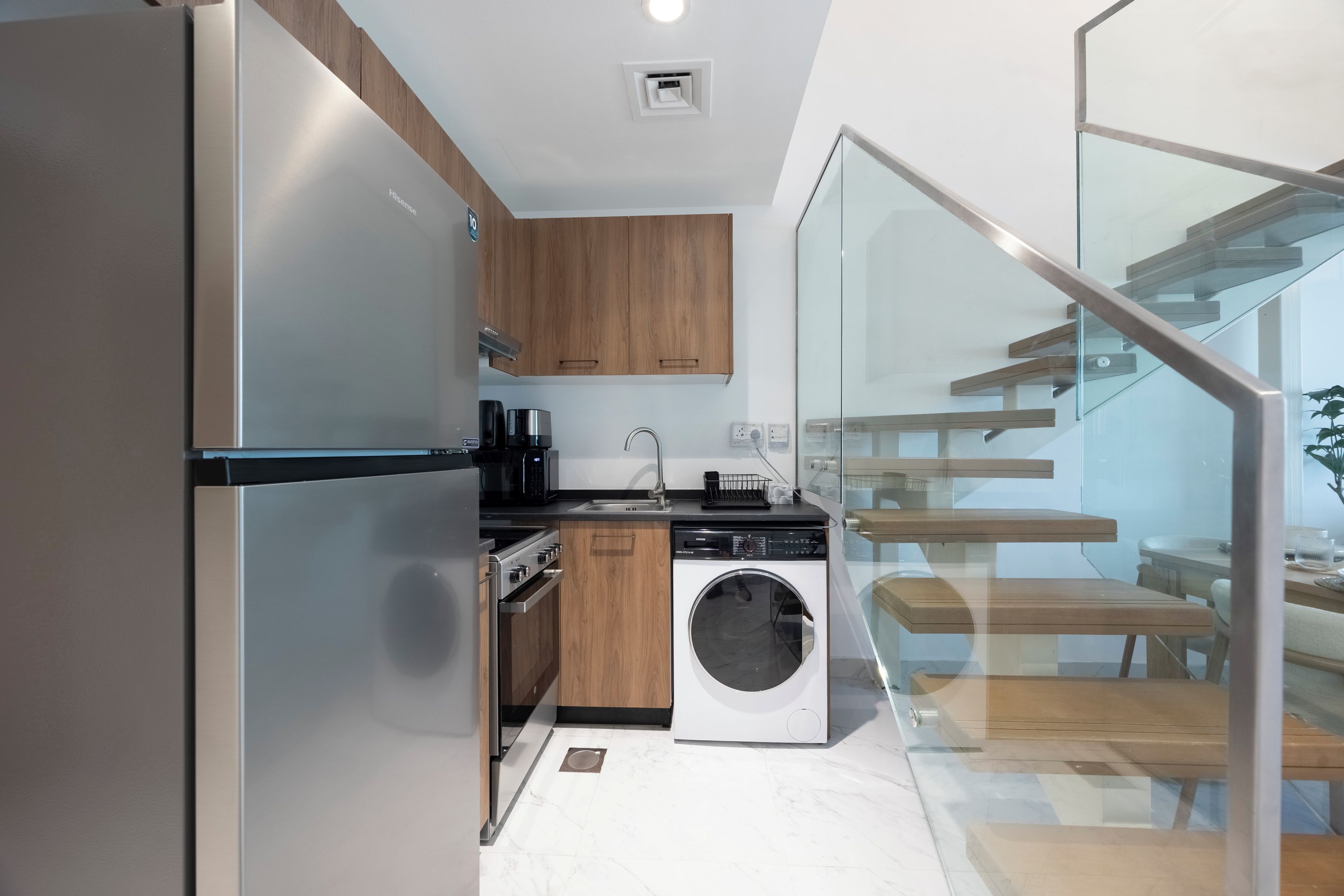 Een moderne keuken met een ingebouwde wasmachine en droger, houten kasten en een glazen trap naar een eetruimte.