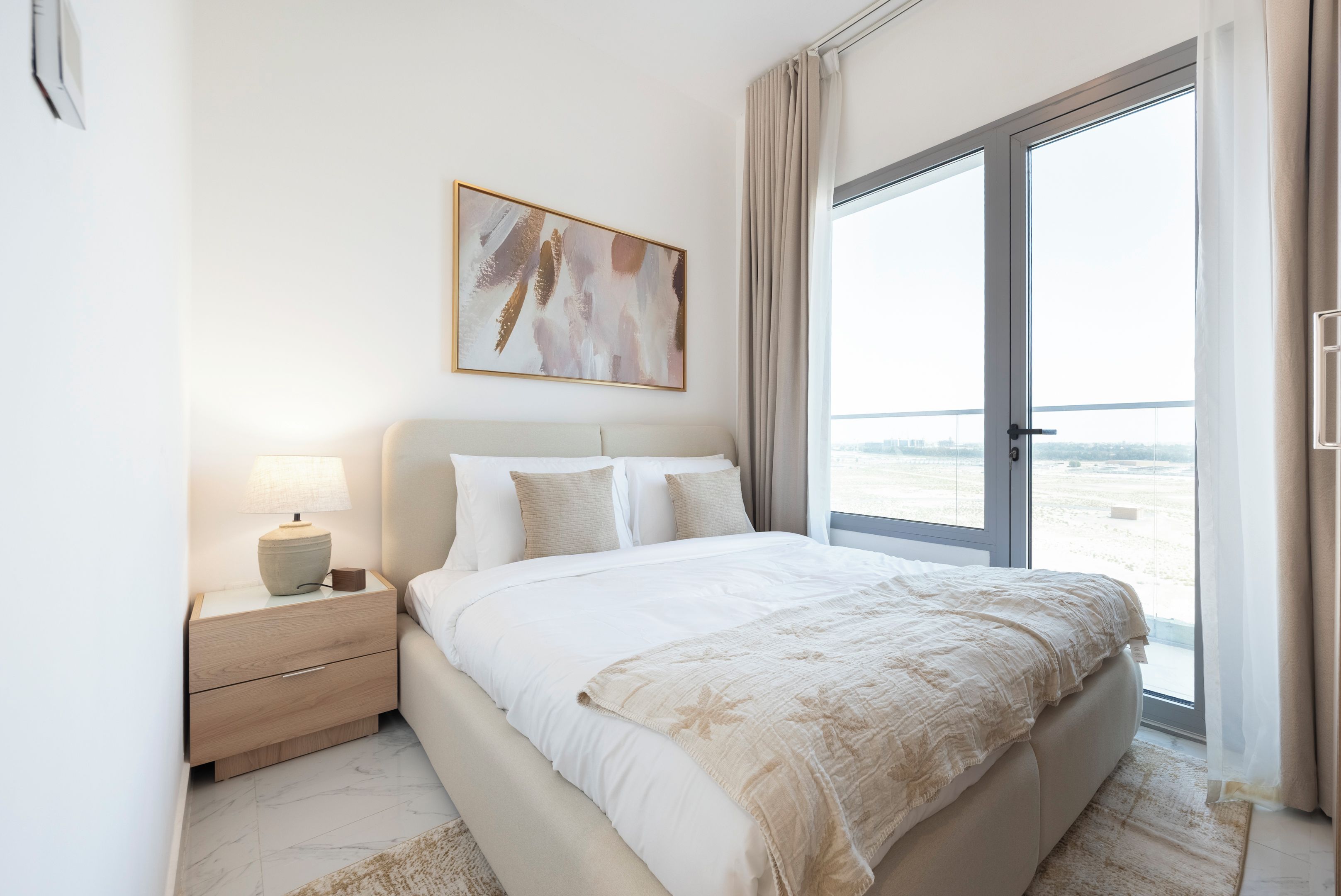 A bedroom with a large bed, a nightstand, a lamp, a painting, and a view through a sliding glass door.