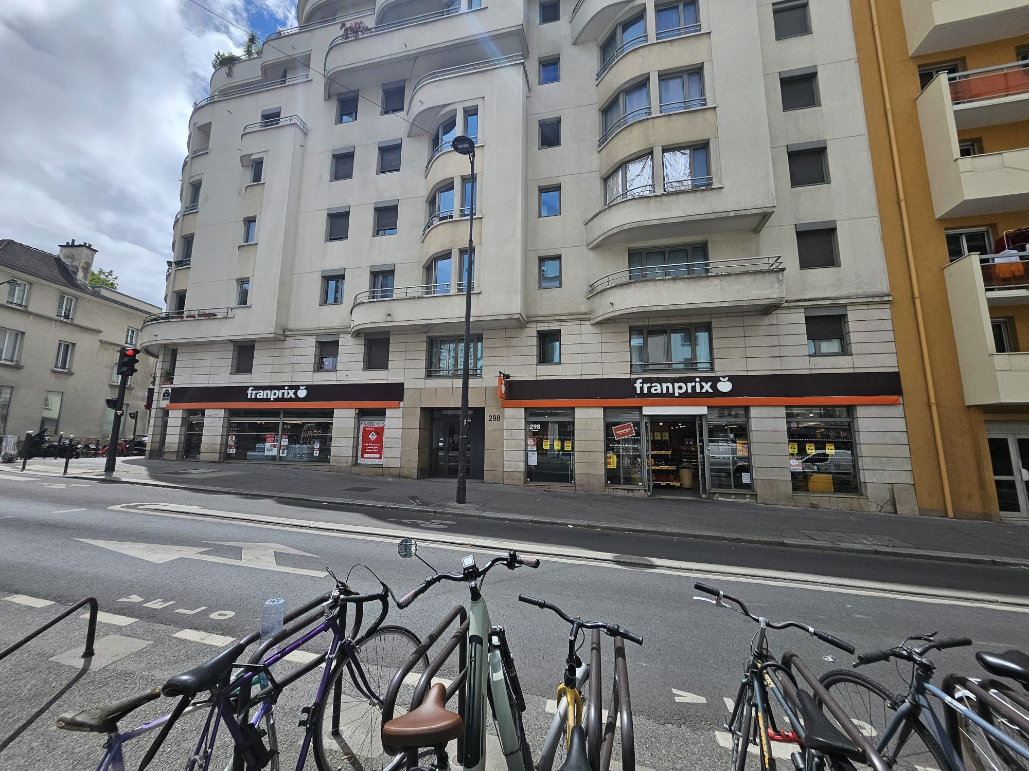 Un immeuble à plusieurs étages, avec un magasin « Franprix » au rez-de-chaussée, un espace de stationnement pour vélos devant et un feu de signalisation.
