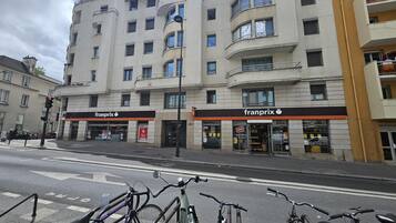 Un immeuble à plusieurs étages, avec un magasin « Franprix » au rez-de-chaussée, un espace de stationnement pour vélos devant et un feu de signalisation.