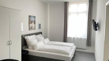 A hotel room with a bed, a wardrobe, a desk, a chair, a window with curtains, and wall-mounted artwork.