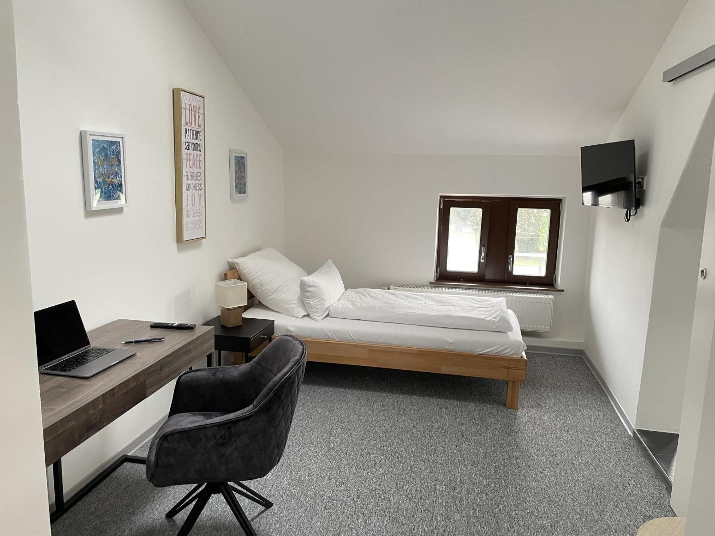 A small, modern room with a bed, a desk with a laptop, a chair, and a TV mounted on the wall.