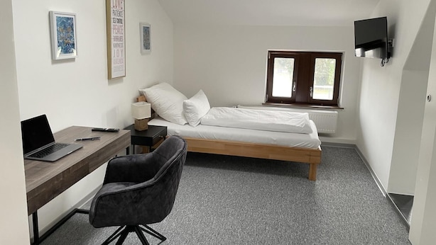 Ein kleiner, moderner Raum mit einem Bett, einem Schreibtisch und einem Stuhl. An der Wand hängen Bilder in Rahmen und über dem Bett ist ein Fernseher angebracht.
