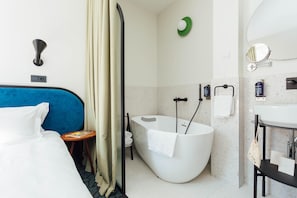 Ett hotellrum med en säng, ett badrum med badkar, en handduksställning och ett litet bord med en tidning.