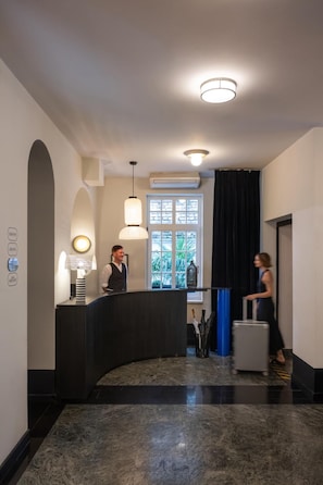 En hotellobby med en böjd receptionsdisk, en man som står bakom den, en kvinna som går förbi och ett stort fönster med utsikt över grönområden.