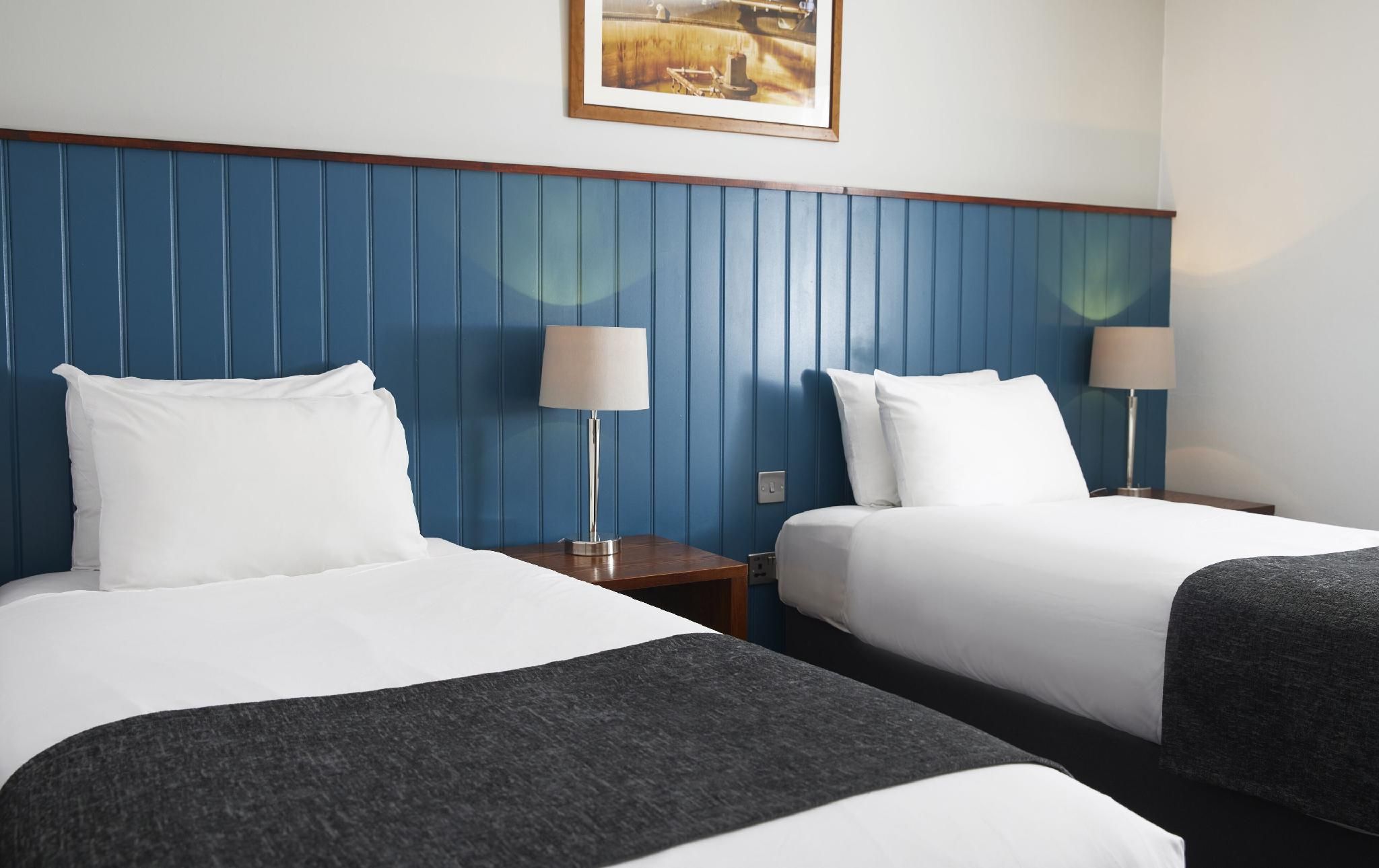 A hotel room with two beds, each with white bedding and a dark throw blanket. There are two bedside tables with lamps, and a framed picture hangs above the beds.