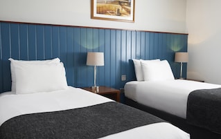Habitación de hotel con dos camas, cada una con ropa de cama blanca y una manta oscura. Hay dos mesitas de noche con lámparas, y un cuadro enmarcado sobre las camas.