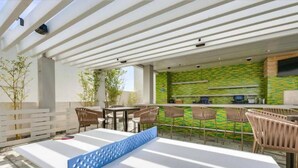 Uma área de varanda externa com mesa branca, cadeiras e um banco azul. Há uma cozinha com painel de fundo verde e um balcão.