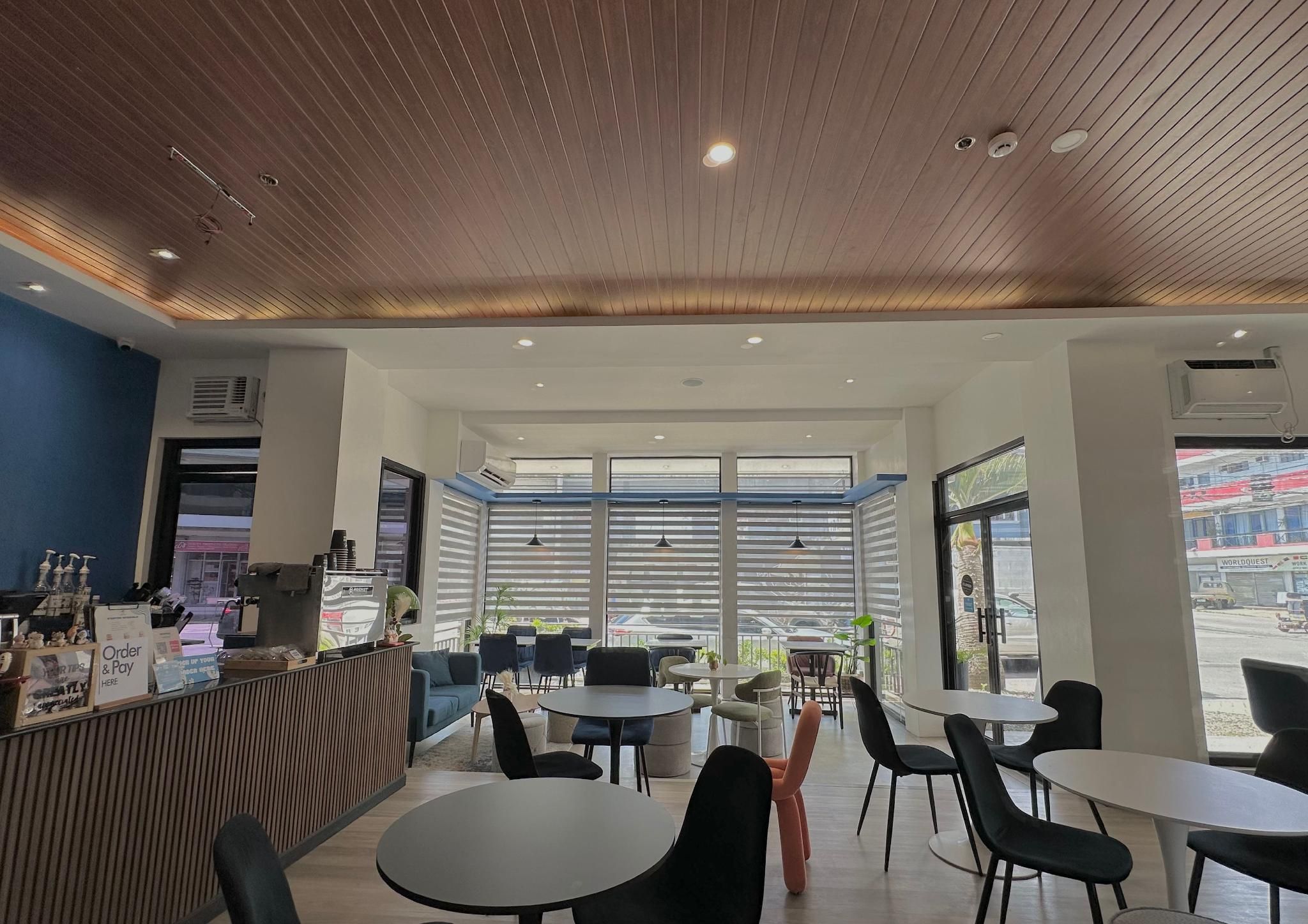 A modern cafe interior with a wooden ceiling, multiple tables and chairs, and large windows offering a view of the street.