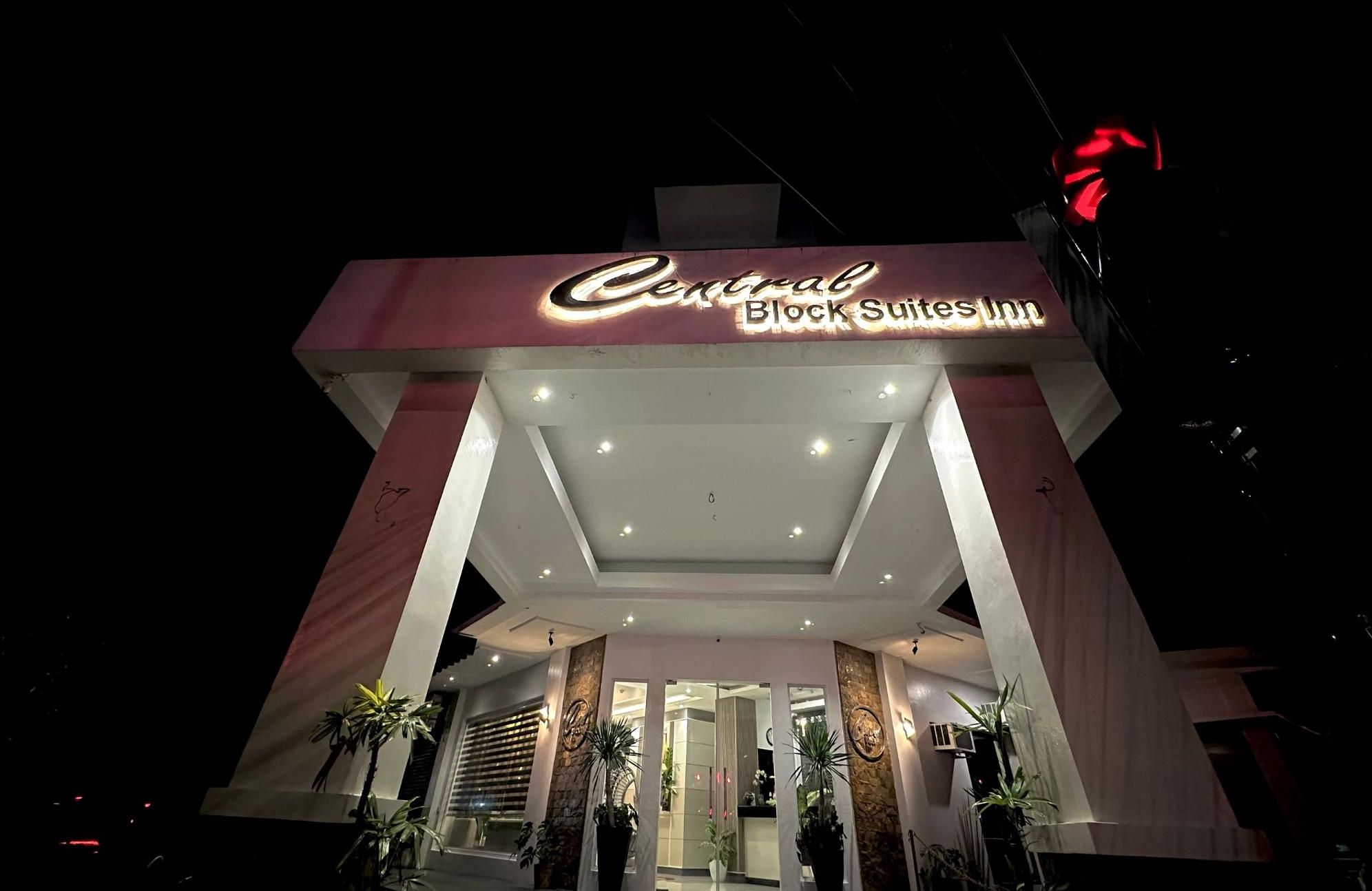 The exterior of Central Block Suites Inn with a prominent illuminated sign, a modern entrance with glass doors, and potted plants at the entrance.