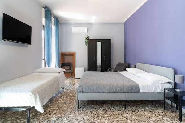 Quarto de hotel com duas camas de solteiro, televisão, guarda-roupa e pequena escrivaninha com luminária.