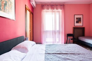 Quarto com uma cama, uma cadeira, uma escrivaninha e uma janela com cortinas leves.