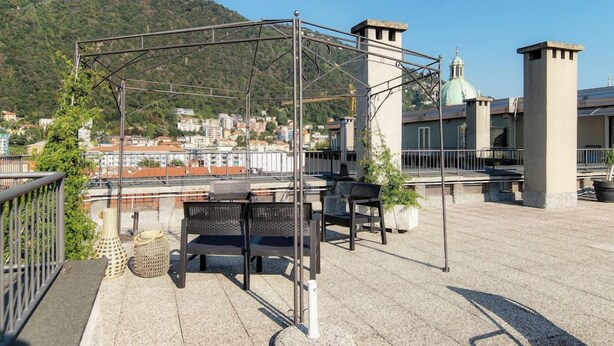 A rooftop terrace with outdoor seating, a pergola, and a view of a cityscape and hill. - Terrazza Rooftop (Como)