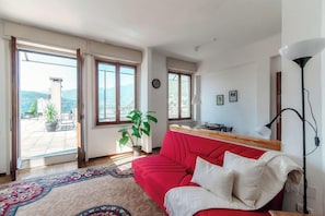 Une pièce avec un canapé rouge, un tapis à motifs, une plante en pot et une vue dégagée sur l’extérieur grâce à de grandes fenêtres.