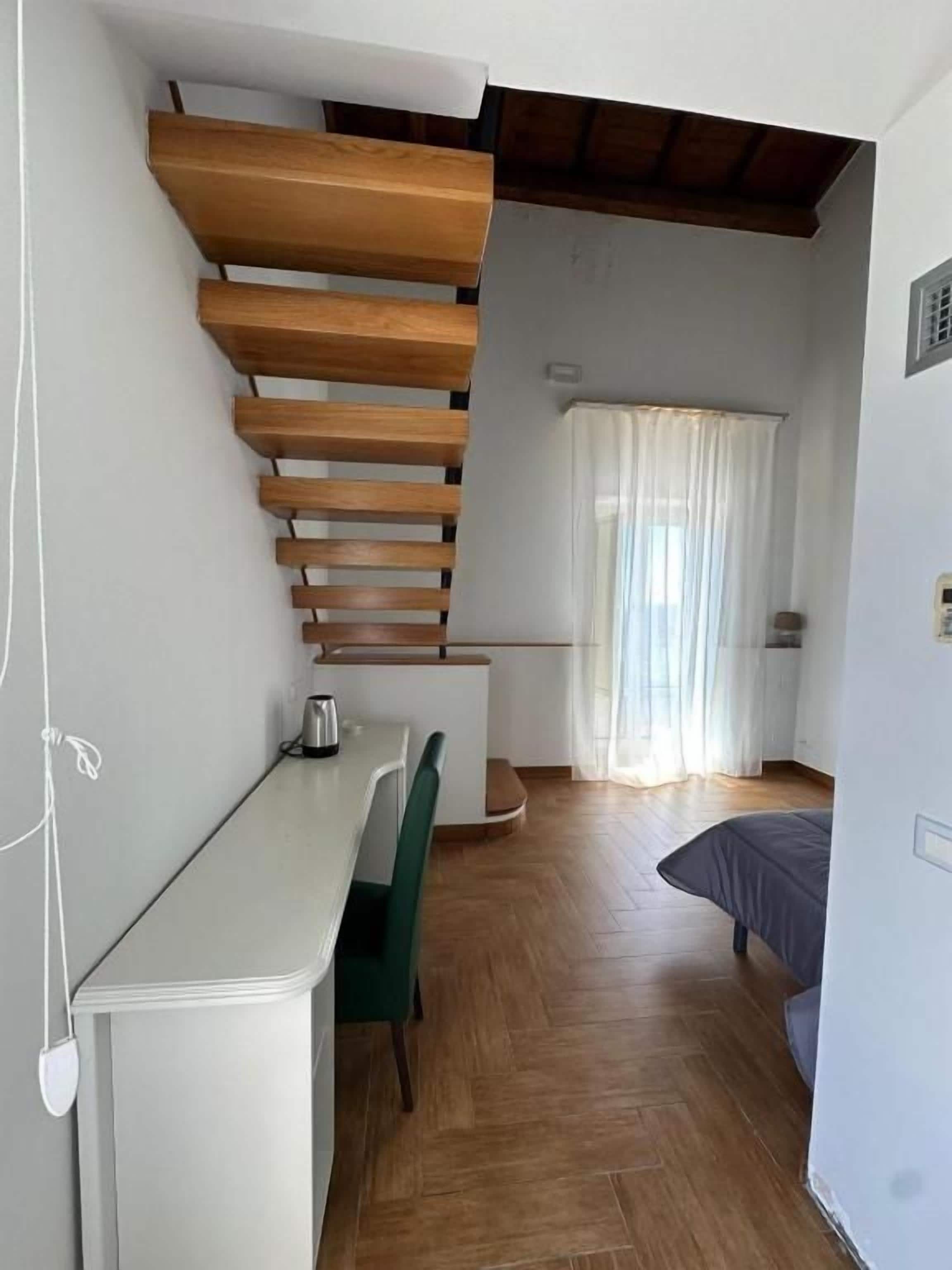 A compact room with a wooden staircase, a small table, a chair, a bed, and a window with curtains.