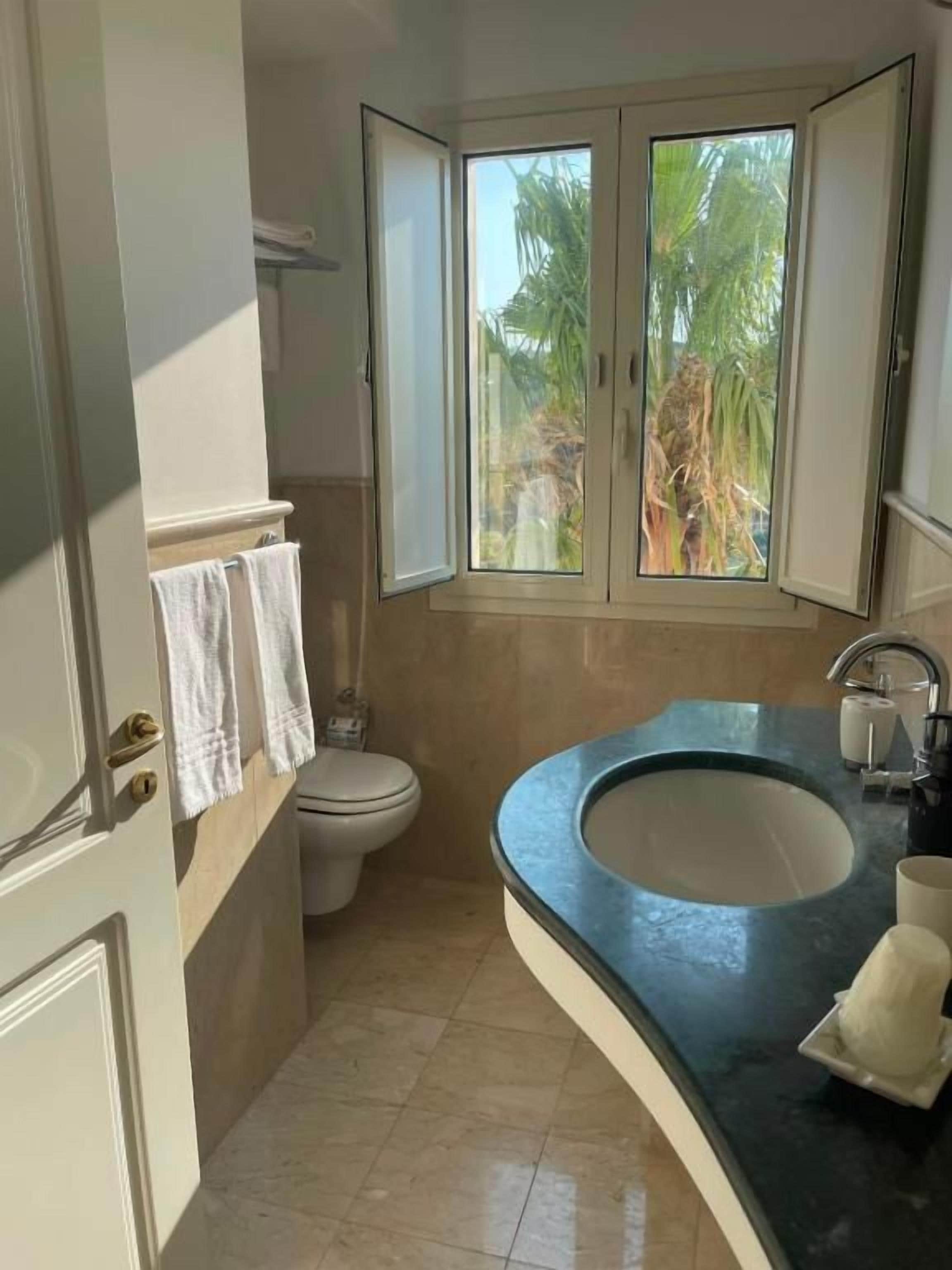 Ein Badezimmer mit Toilette, Waschbecken und einem Fenster mit Blick auf Palmen.