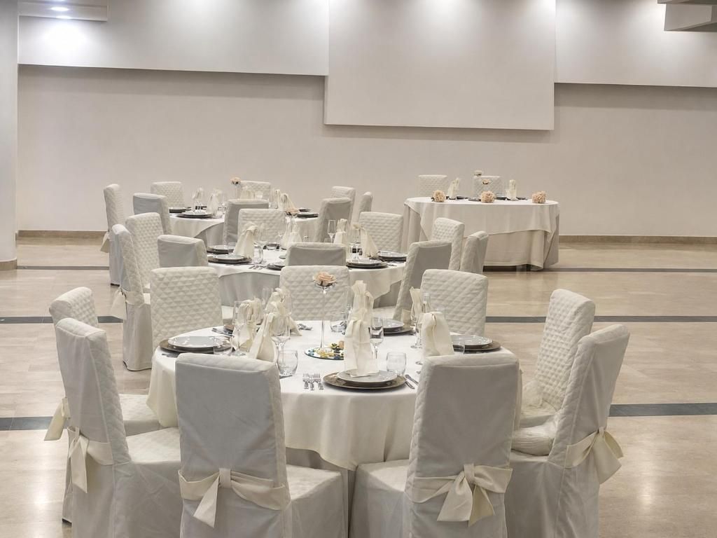 Un salón de banquetes con mesas redondas dispuestas para cenar, sillas blancas y una decoración en tonos neutros.