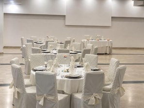 Uma sala de banquetes com mesas redondas preparadas para refeições, cadeiras brancas e uma paleta de cores neutras.