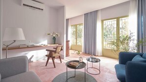 A modern living room with a sofa, a chair, a coffee table, and a desk with a lamp. There is a window with curtains and a view of greenery outside. - M Village Living Dien Bien Phu (Ho Chi Minh Municipality)