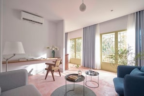 A modern living room with a sofa, a chair, a coffee table, and a desk with a lamp. There is a window with curtains and a view of greenery outside. - M Village Living Dien Bien Phu (Ho Chi Minh Municipality)