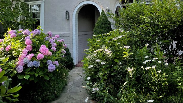Una casa amb teulada inclinada, un camí de pedra i diverses plantes amb flors.