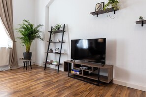 Ein modernes Wohnzimmer mit einem Flachbildfernseher auf einem Holzständer, einer Zimmerpflanze, einem Hängeregal und einem kleinen Regal mit Dekorationsobjekten.