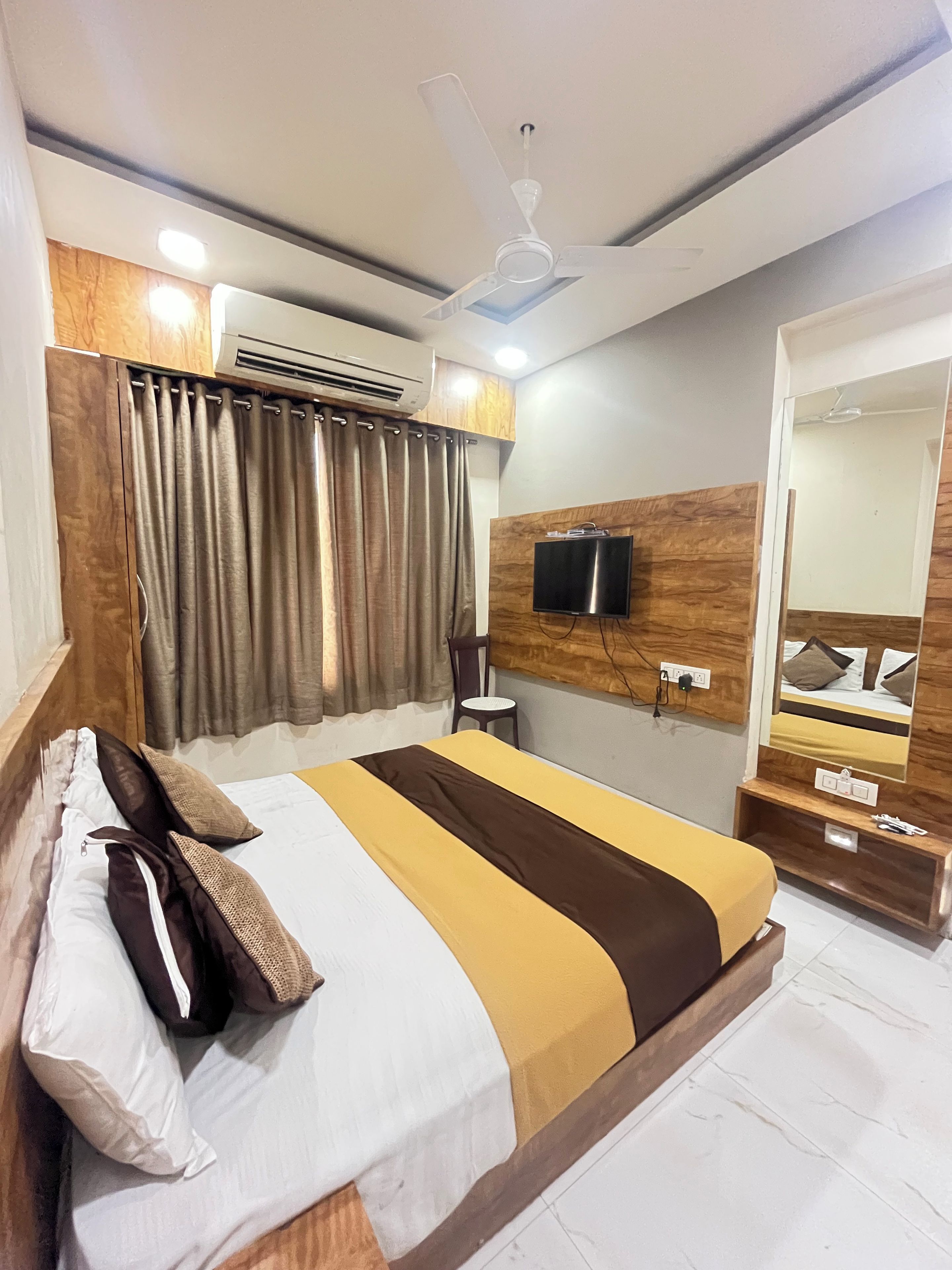 Habitación de hotel con cama, cortinas, aire acondicionado, ventilador de techo, televisor y espejo.