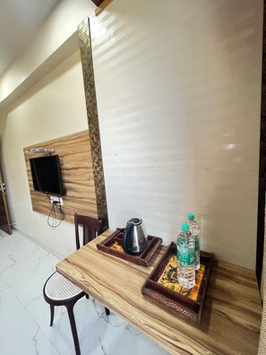 A dining area with a wooden table, a chair, a television mounted on the wall, and a tray with a kettle and water bottles.