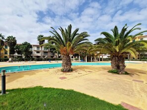 A pool area with palm trees, a clear sky, and a building in the background. - Sunny 1-Bedroom Apartment Island Retreat with Pool Views at Vila Verde Resort (Sal)