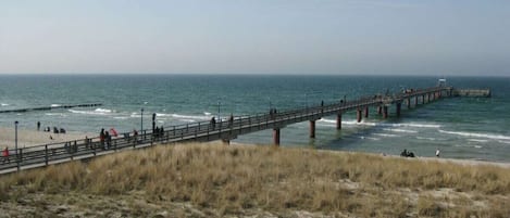 Escena costera con un muelle que se adentra en el mar, playa arenosa y dunas cubiertas de hierba.