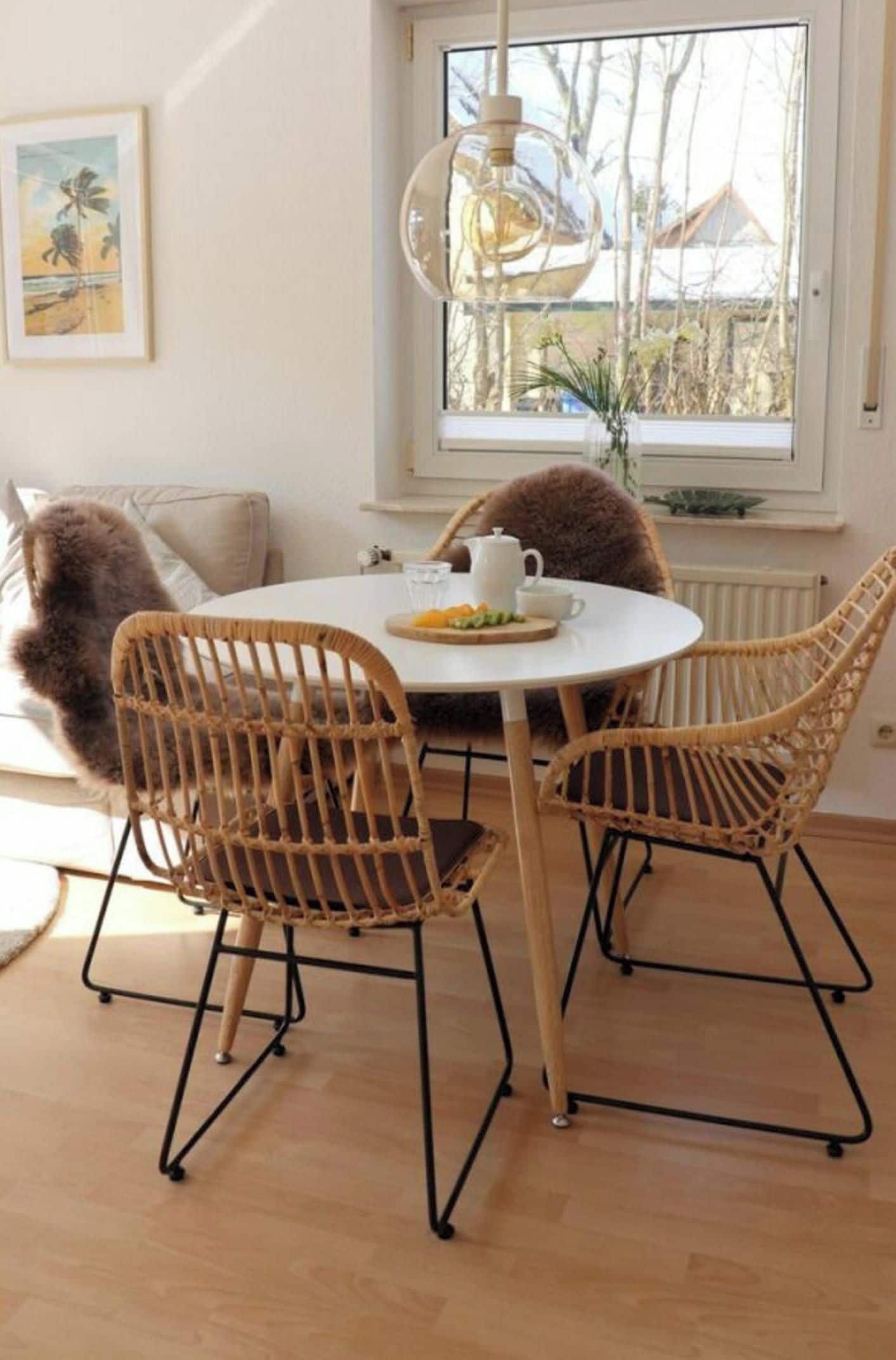 A dining area with a round table, wicker chairs, a hanging light fixture, and a window with a view of trees and buildings.
