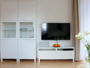 A modern living room with a flat-screen TV mounted on a white cabinet, a glass shelf with dishes, and a vase with flowers on a table. - Holiday Apartment With 2 Bedrooms (Zingst)