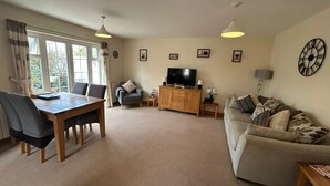 A living room with a dining table, sofa, television, and a clock on the wall. - Guernsey 2 bedroom cottage near Perelle Bay (Saint Saviour)