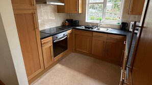 A kitchen with wooden cabinets, a stainless steel range hood, a built-in oven, a sink, and a window with a view of greenery. - Guernsey 2 bedroom cottage near Perelle Bay (Saint Saviour)