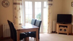 A dining area with a table and chairs, a clock on the wall, a television on a stand, and a window with curtains. - Guernsey 2 bedroom cottage near Perelle Bay (Saint Saviour)