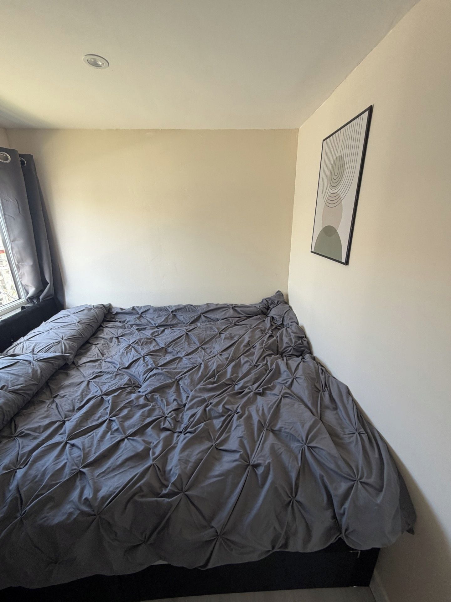 A single bed with a grey duvet in a room with a window, a picture on the wall, and a ceiling light.