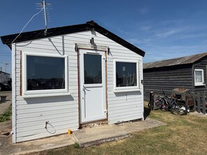 A small, single-story building with a door and two windows, situated next to a wooden fence and a bicycle. - Cabin 161 Sleeps 7 With Garden and Parking (Sheerness)