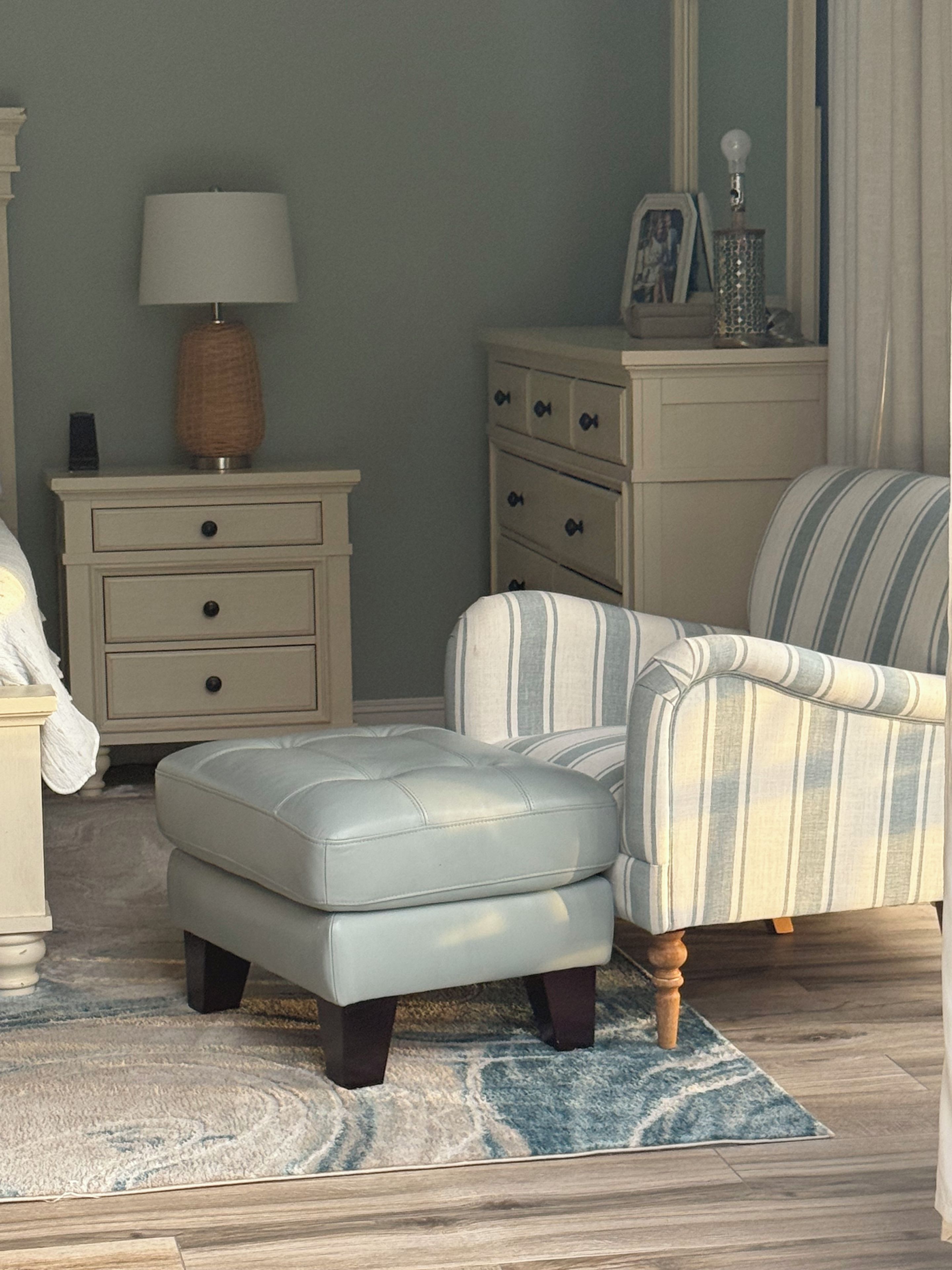 A bedroom with a bed, a nightstand, a lamp, a dresser, a chair, an ottoman, and a rug.