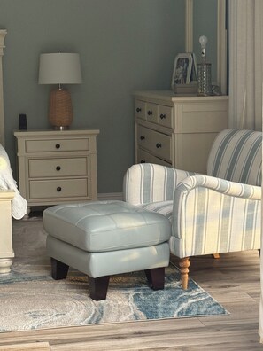 Een slaapkamer met een bed, nachtkastje, lamp, dressoir, stoel, poef en vloerkleed.
