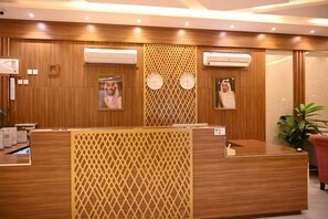 A reception desk with a wooden finish, air conditioning units, framed pictures on the wall, and a potted plant. - Jawharat Al Sarat for hotel apartments (Al-Amjad District)