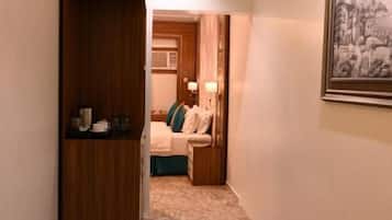 A hallway leading to a bedroom with a bed, a bedside table, and a nightstand. There is a framed picture on the wall.