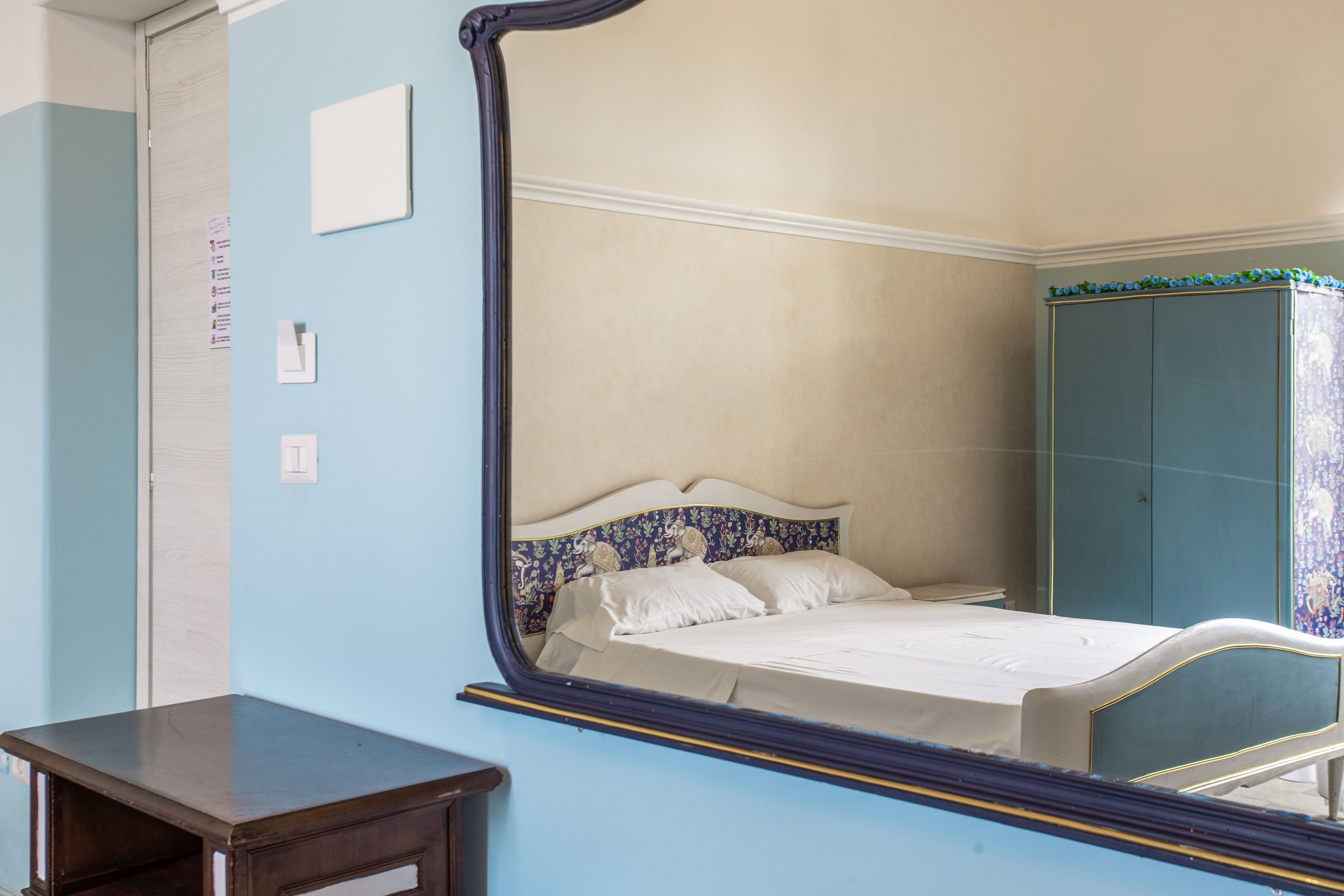 A bedroom with a large mirror reflecting the bed and wardrobe, a small bedside table, and a door with a light switch.