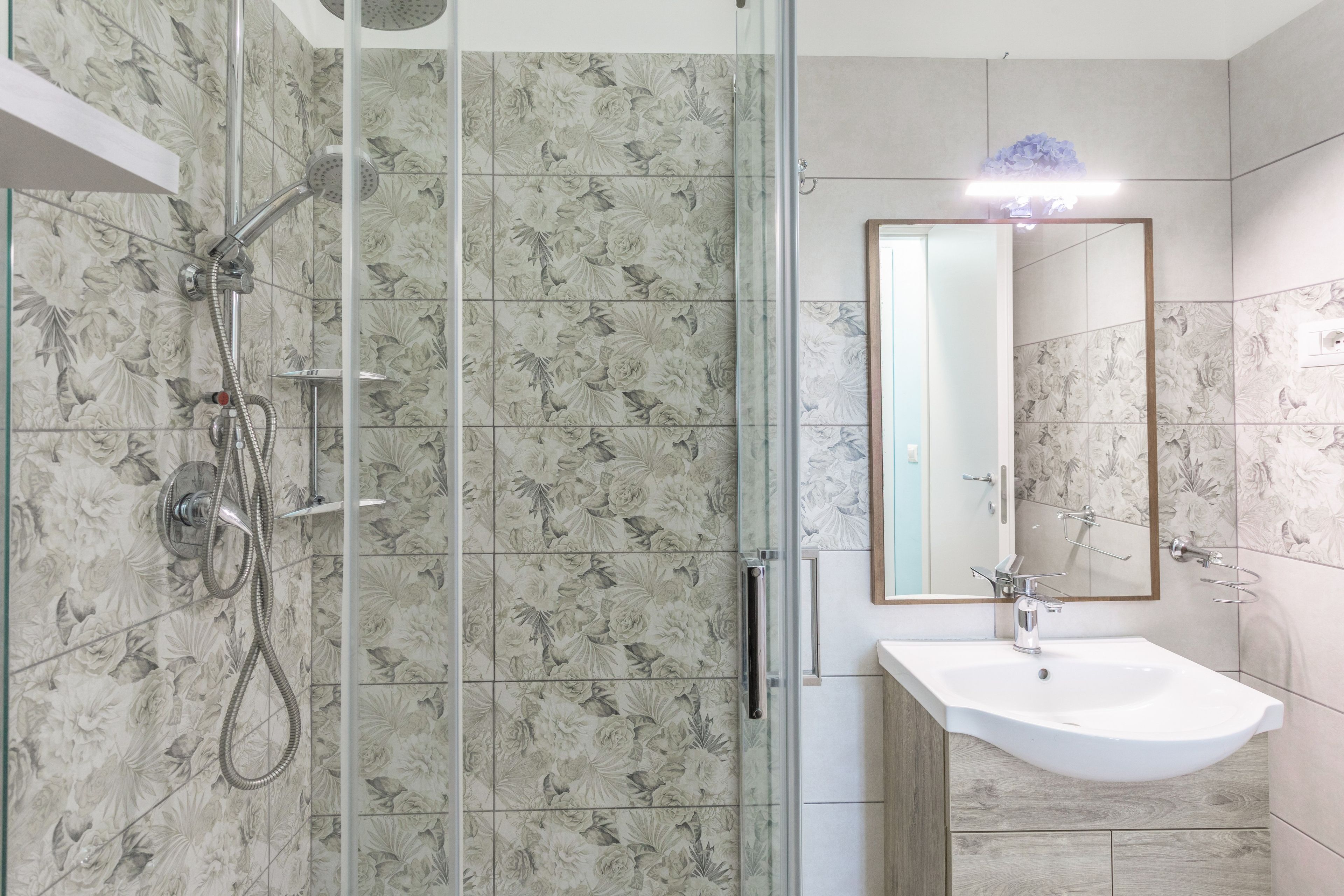 Un bany modern amb plat de dutxa envoltat de vidre, un lavabo amb mirall i rajoles amb motius florals.