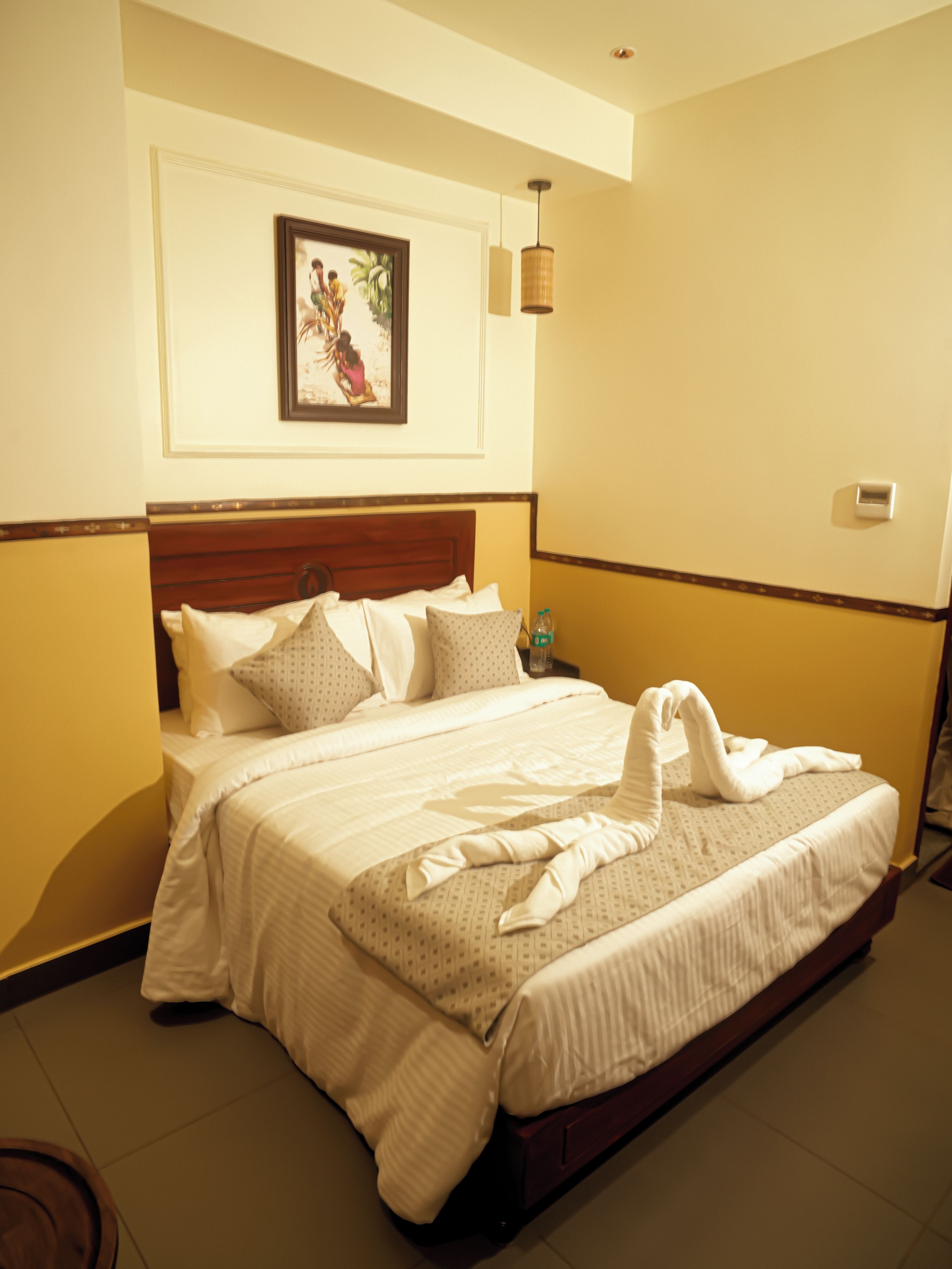 A hotel room with a bed, pillows, a framed picture on the wall, and a bedside table with a bottle.