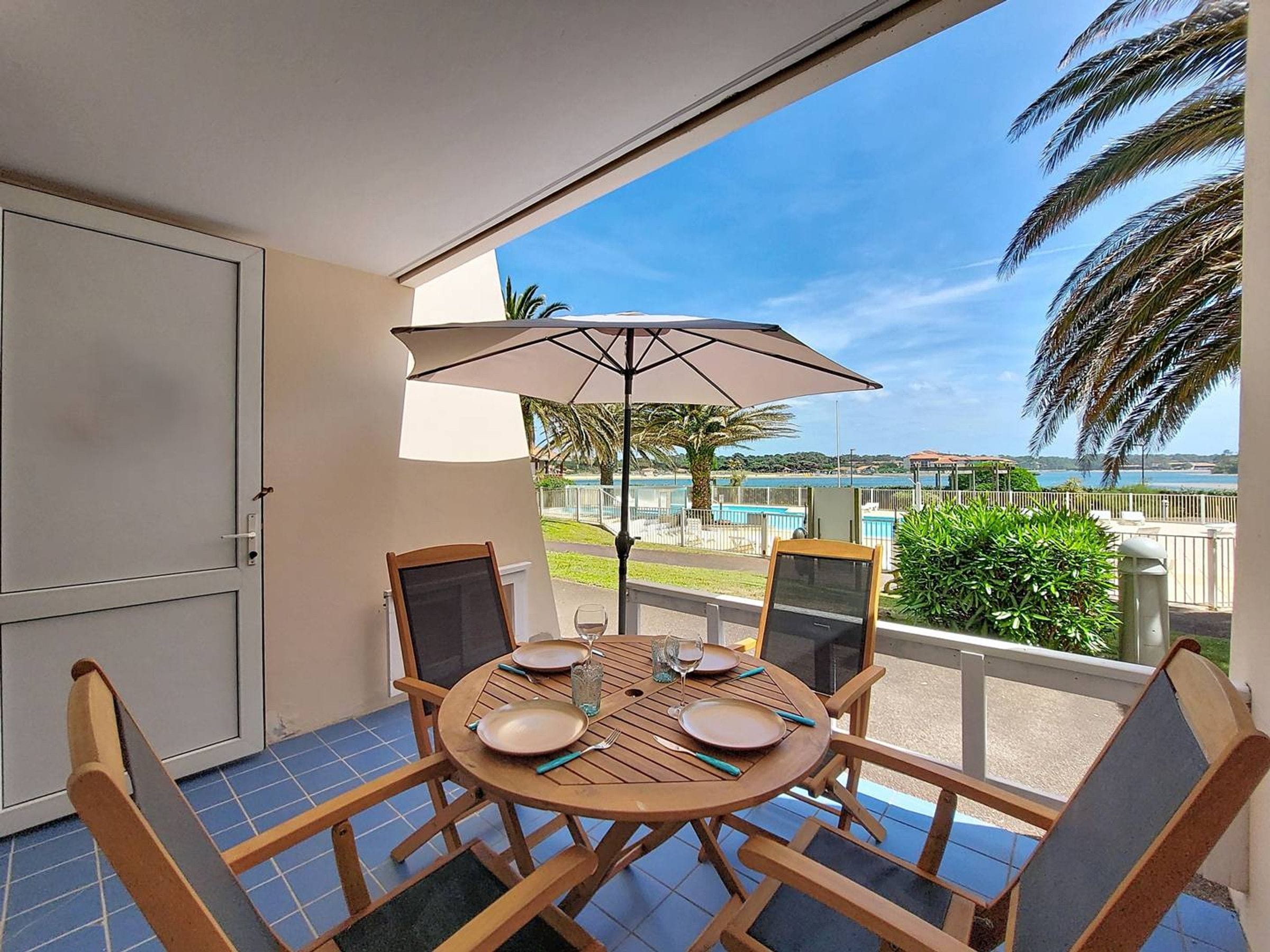 Un balcón con una mesa lista para dos personas, con vista a una piscina y palmeras.