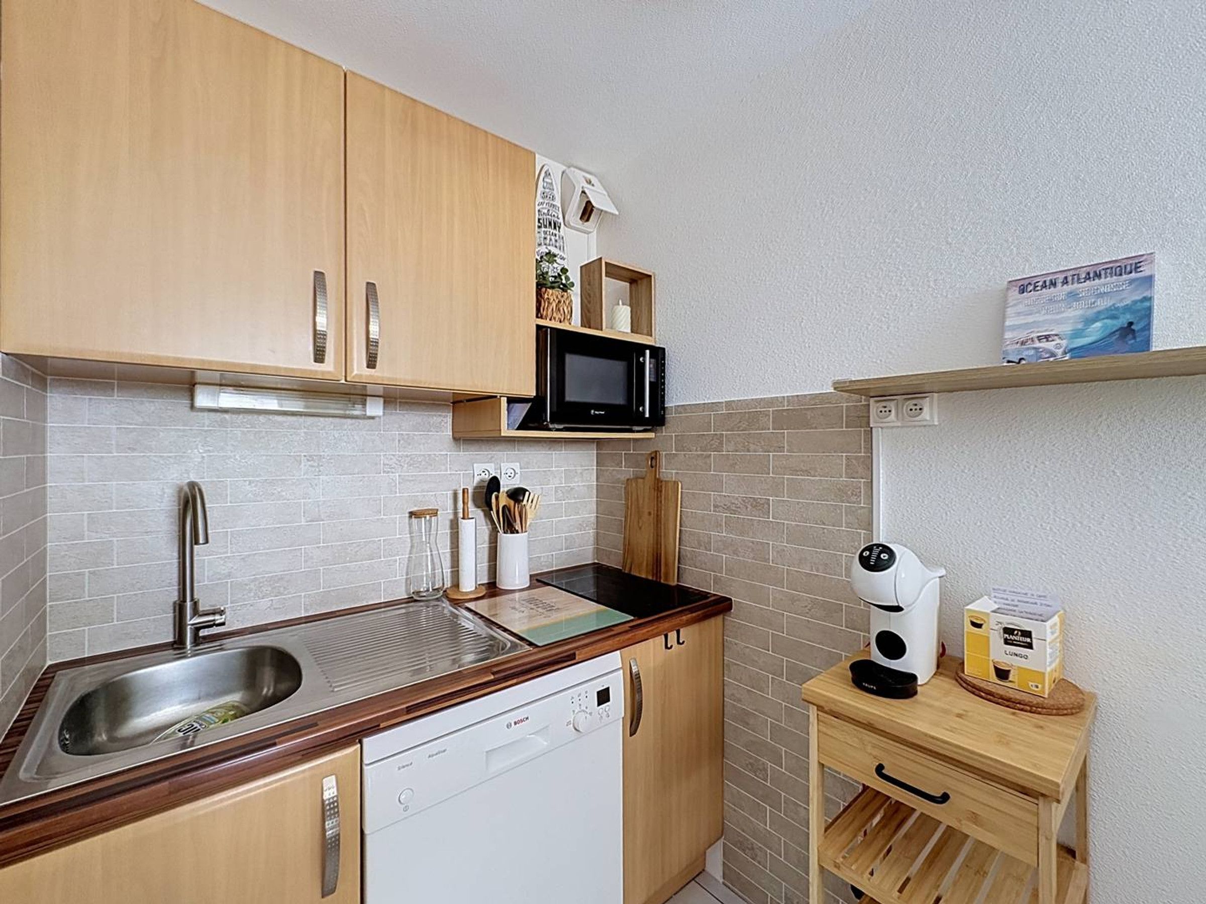 A compact kitchen with wooden cabinets, a stainless steel sink, a Bosch dishwasher, a microwave, and a coffee maker on a small wooden table.