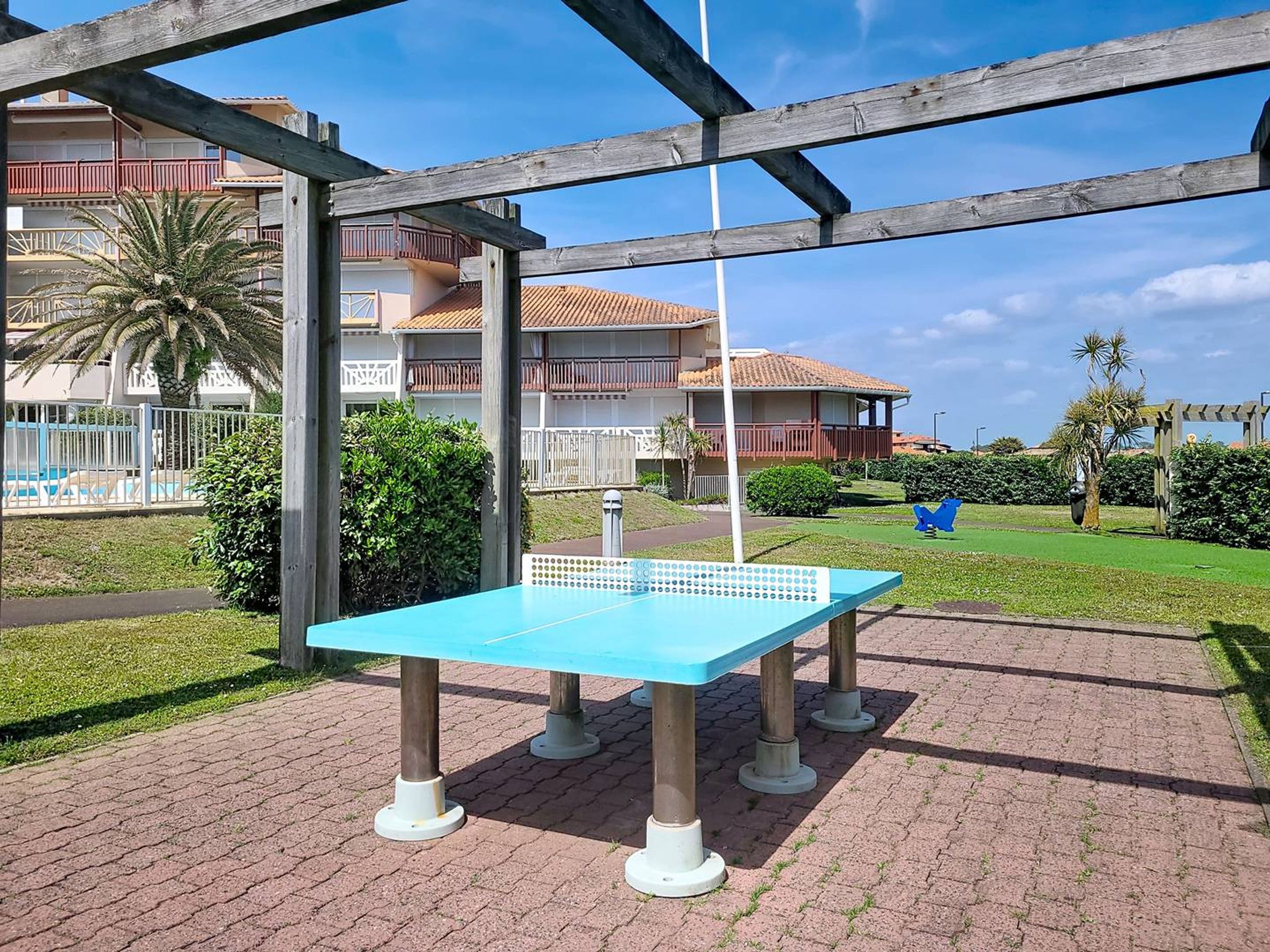 A blue ping pong table in an outdoor area with a brick floor, surrounded by greenery and a building with balconies in the background.