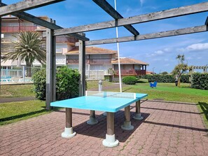 A blue ping pong table in an outdoor area with a brick floor, surrounded by greenery and a building with balconies in the background. - Studio with balcony - 4 guests (Vieux-Boucau-les-Bains)