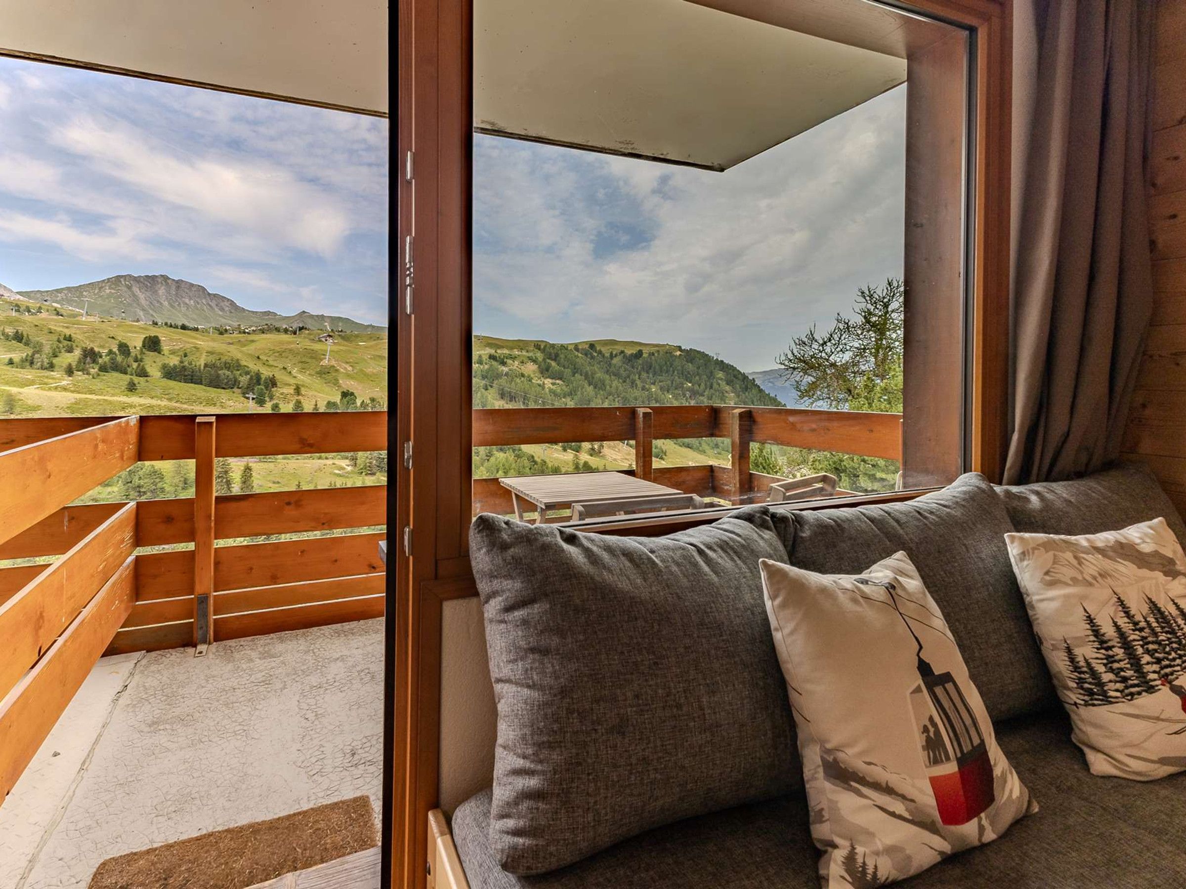 A balcony with a wooden railing, a sofa with cushions, and a view of a mountainous landscape.