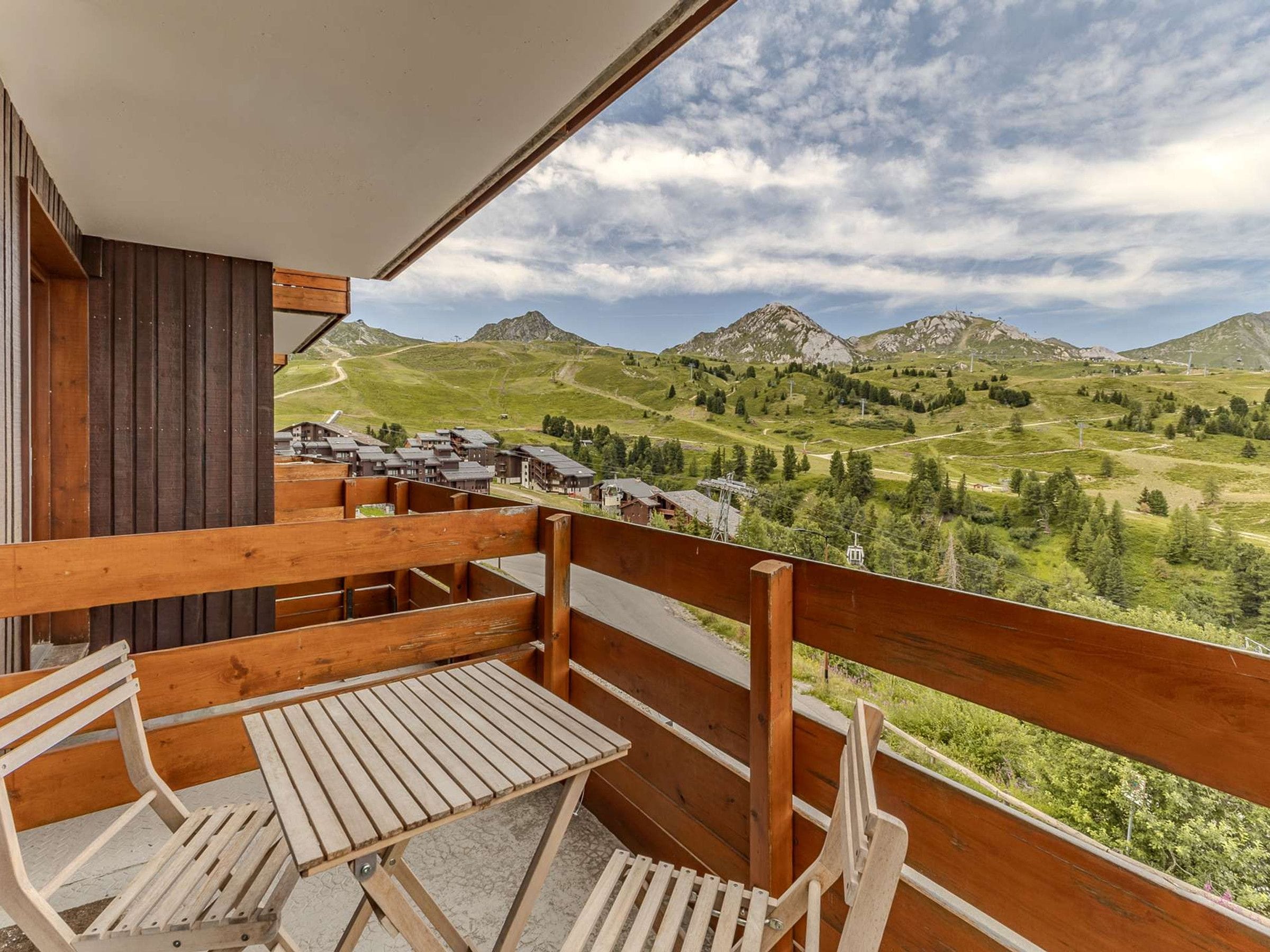 Un balcón de madera con una mesa y sillas, con vistas a un paisaje montañoso con edificios.